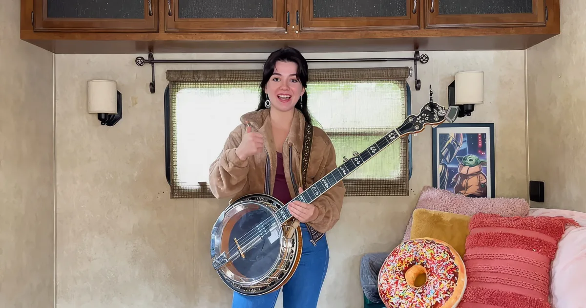A woman in a tan jacket plays a banjo in a cozy RV interior with a window and wall art.