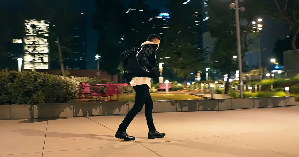 A man in a black jacket and white hoodie walks on a city sidewalk at night, surrounded by illuminated buildings and greenery.