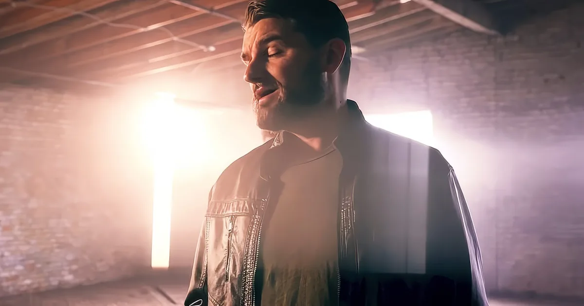 A man with a beard and short hair stands in a dimly lit room with brick walls, wearing a jacket.