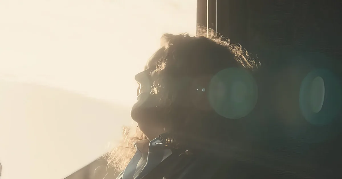 A person with curly hair, wearing a dark jacket, is silhouetted against a bright, hazy background with lens flare.