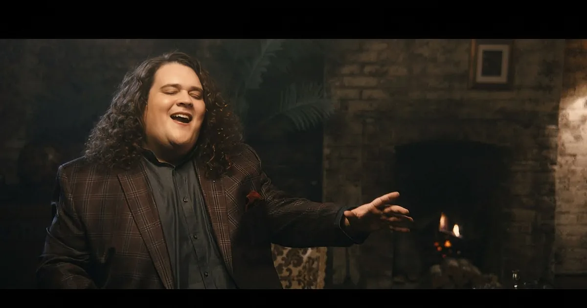 A person with curly hair, wearing a plaid blazer, is smiling and clapping their hands in front of a stone wall.