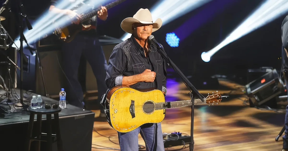 A man in a cowboy hat plays a guitar on stage with bright lights and other musicians.