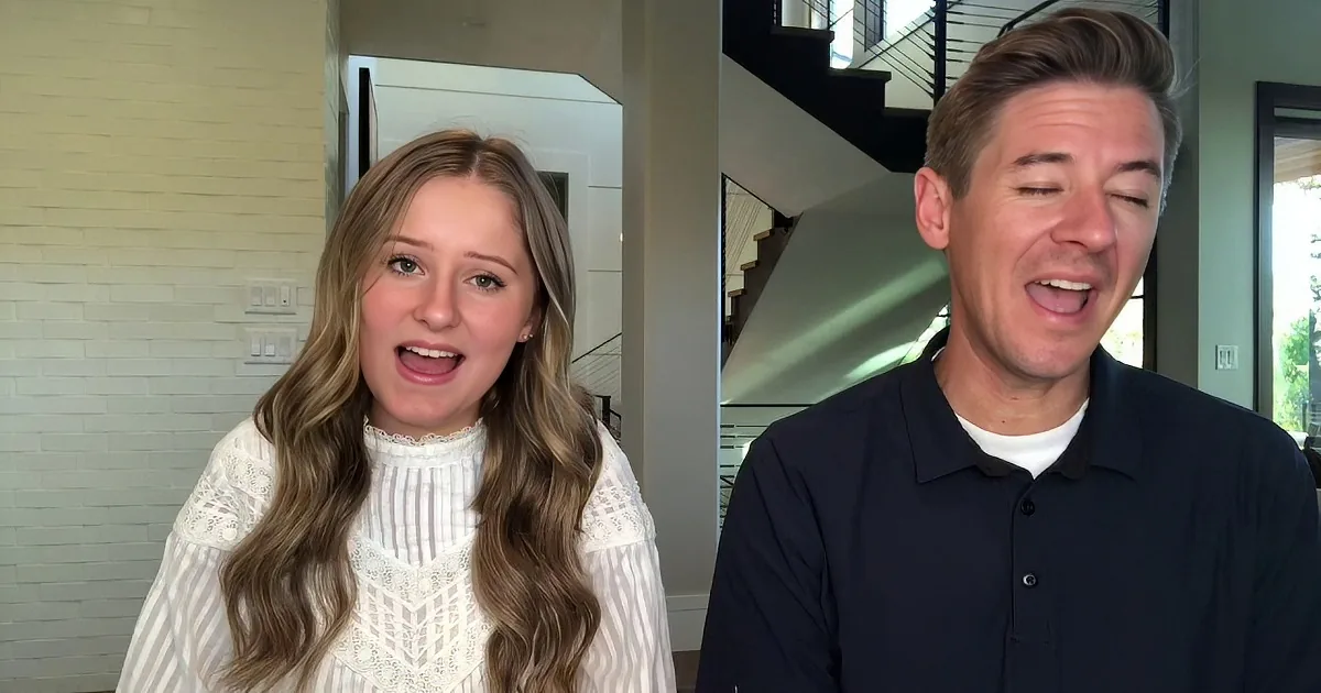 A woman in a white lace top and a man in a dark polo shirt are speaking in a modern home setting.