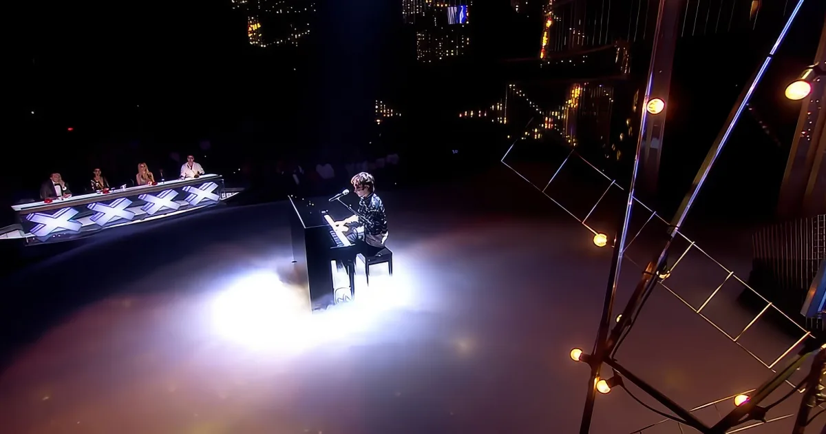 A person plays piano on a dimly lit stage with judges seated at a table in the background.