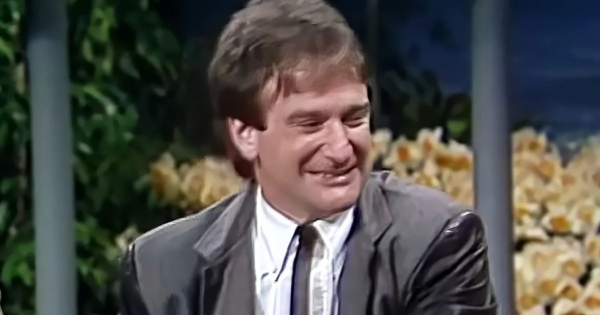 A man in a suit smiles widely, surrounded by blurred greenery and flowers.