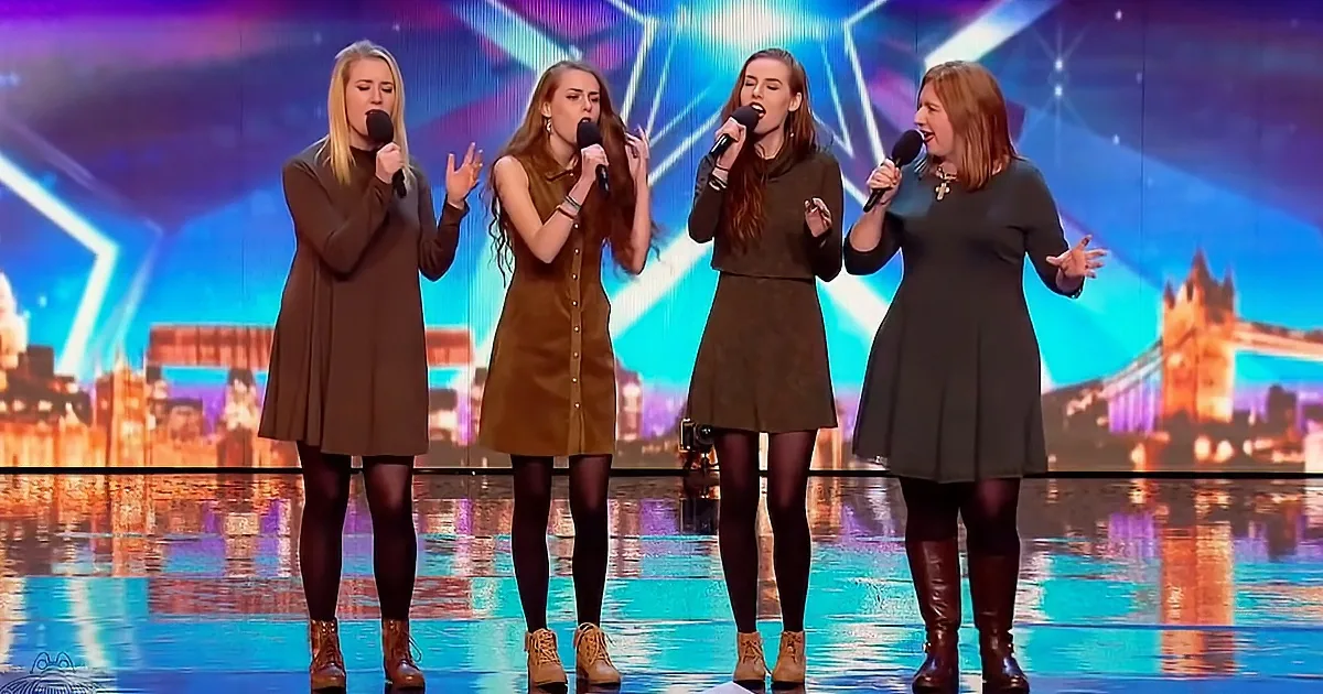 Four women in brown dresses sing on stage with microphones, cityscape backdrop.