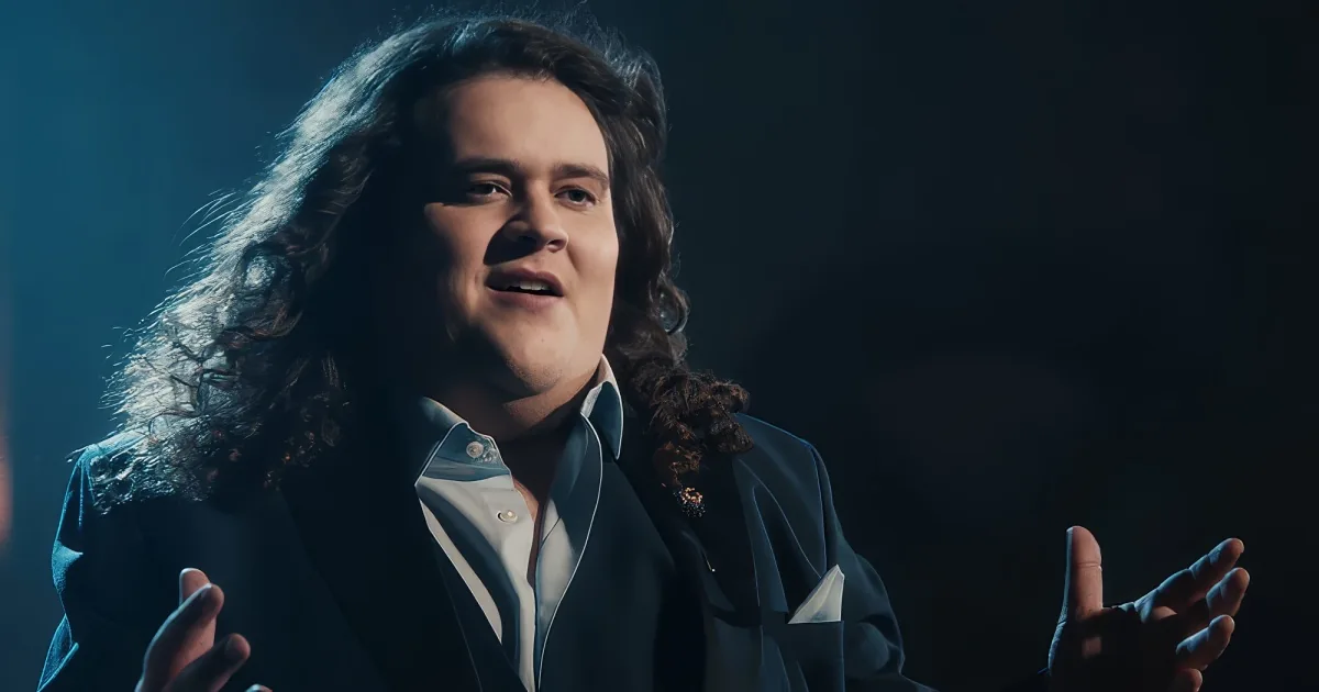A person with long, curly hair and a white shirt is singing passionately with their hands raised.