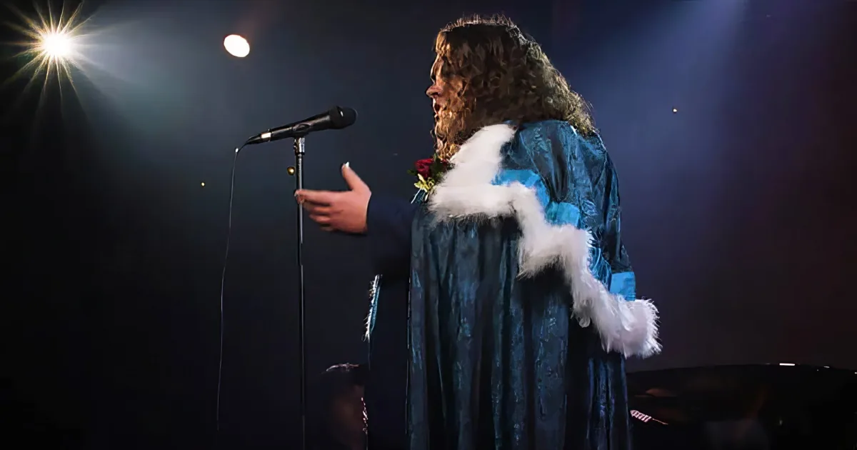 A person in a blue robe with white fur trim stands on stage, gesturing near a microphone.