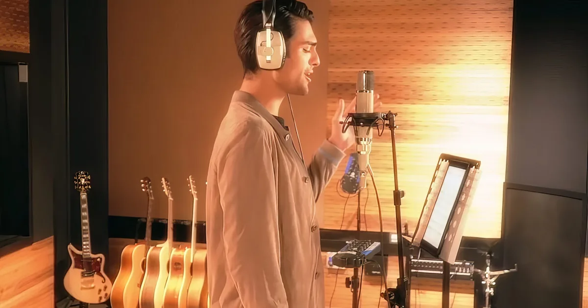 A man wearing headphones sings into a microphone in a recording studio with guitars and equipment.