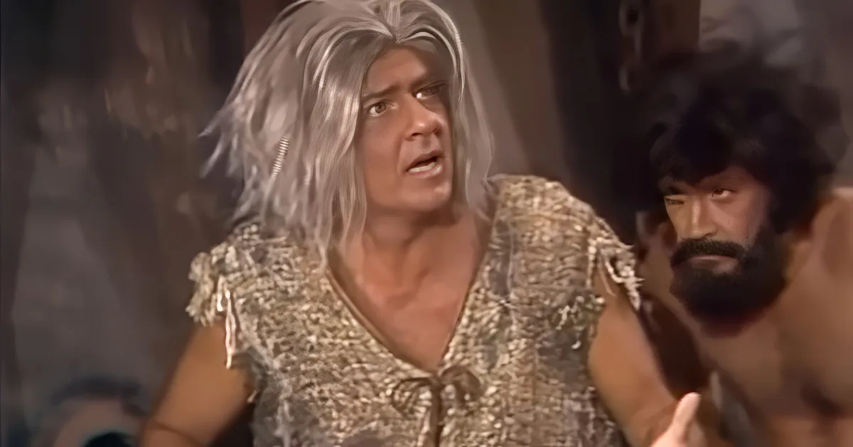 A man with long white hair and a golden outfit is speaking, with another man in a dark outfit beside him.