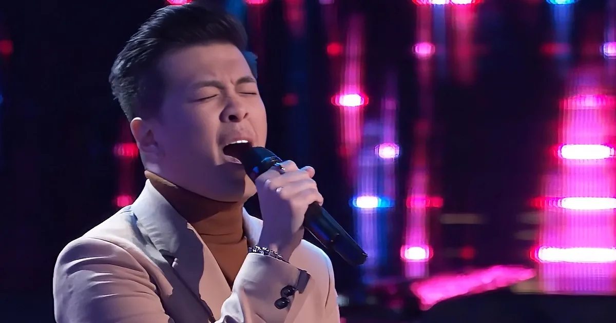 A man in a white suit sings passionately into a microphone on stage with vibrant pink and blue lights.