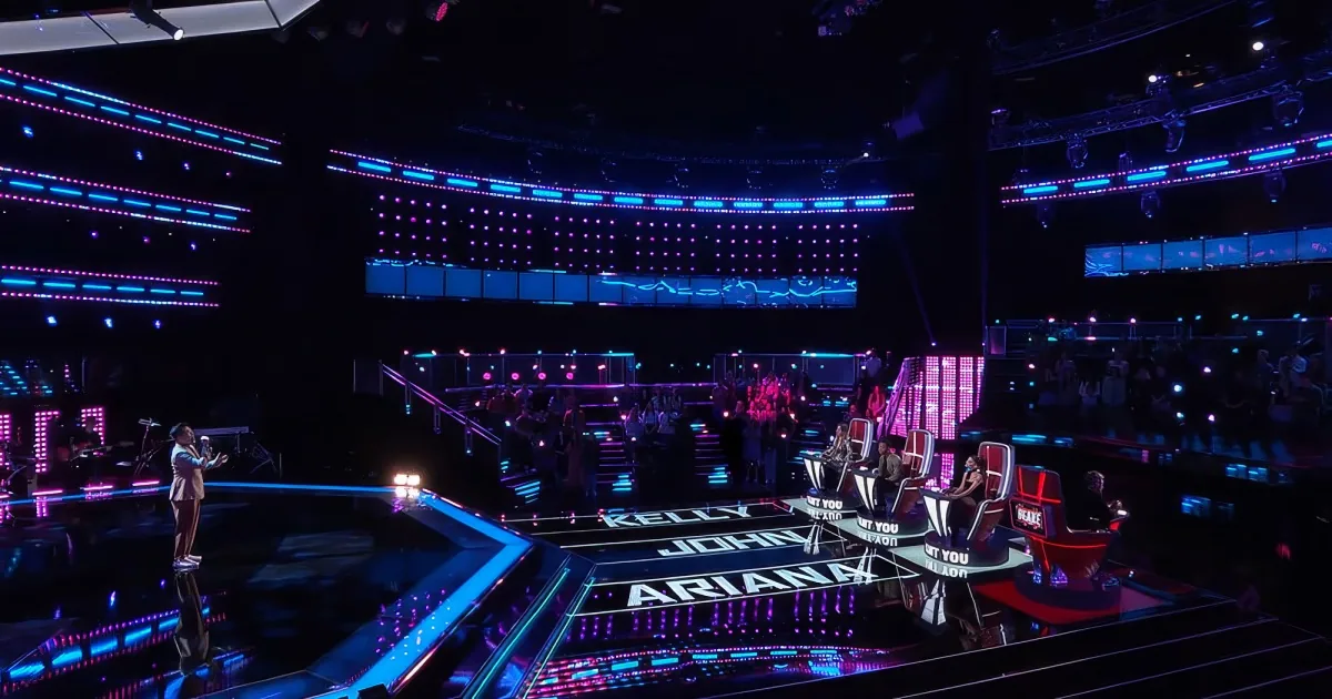 A stage with vibrant purple and blue lighting, featuring judges' chairs and a large audience in a darkened arena.