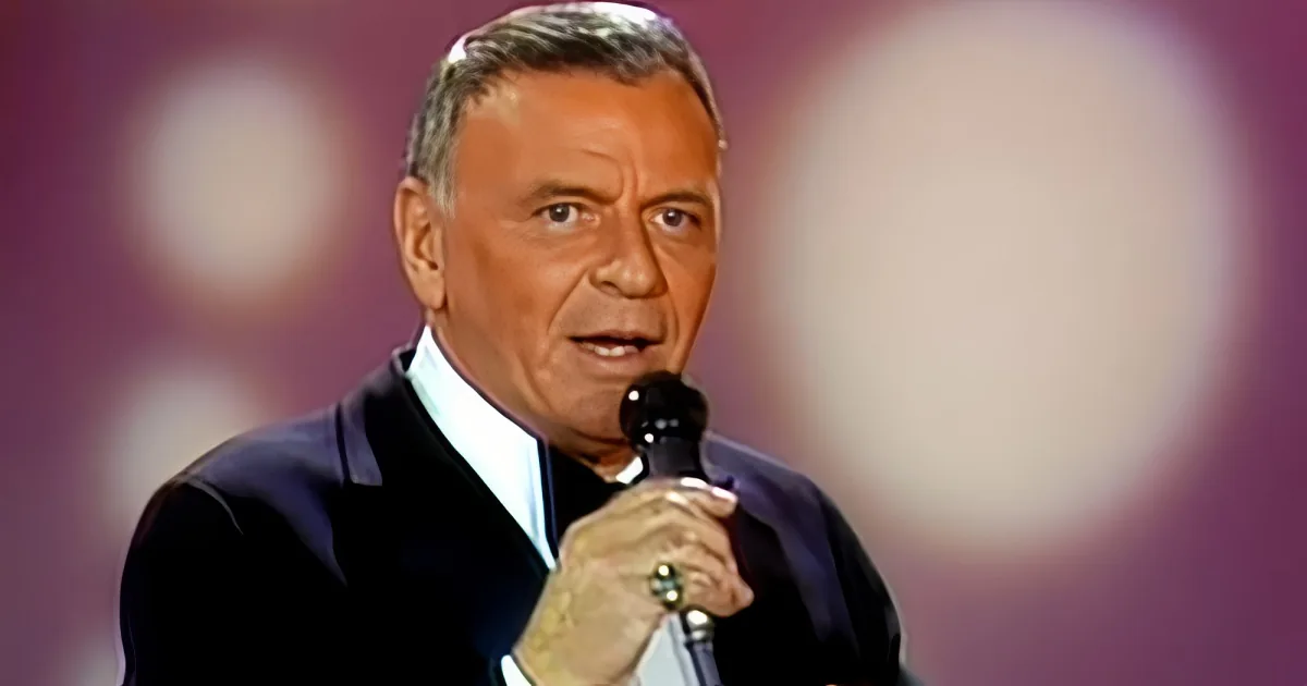 A man in a suit holding a microphone, speaking into it, with a blurred background.