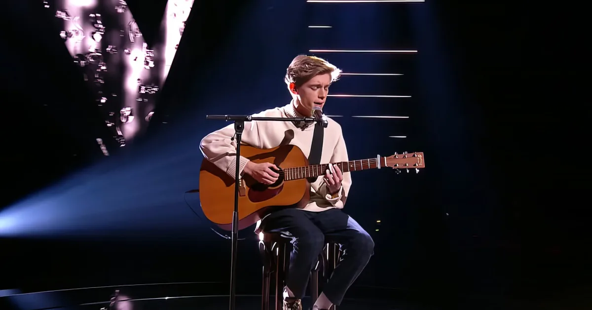 A person plays guitar on stage with a microphone, illuminated by stage lights.