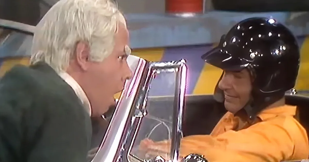 A man in a green suit is talking to another man wearing a black helmet in a car.