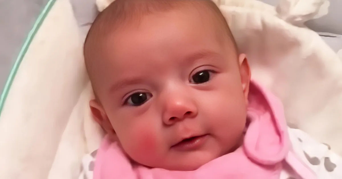 A baby wrapped in pink and white, lying on a white blanket, looking curious.