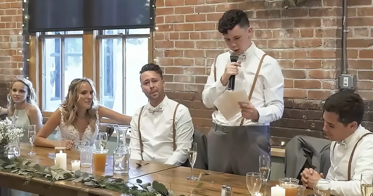A man in white speaks into a microphone at a wedding reception, surrounded by guests at a long table.