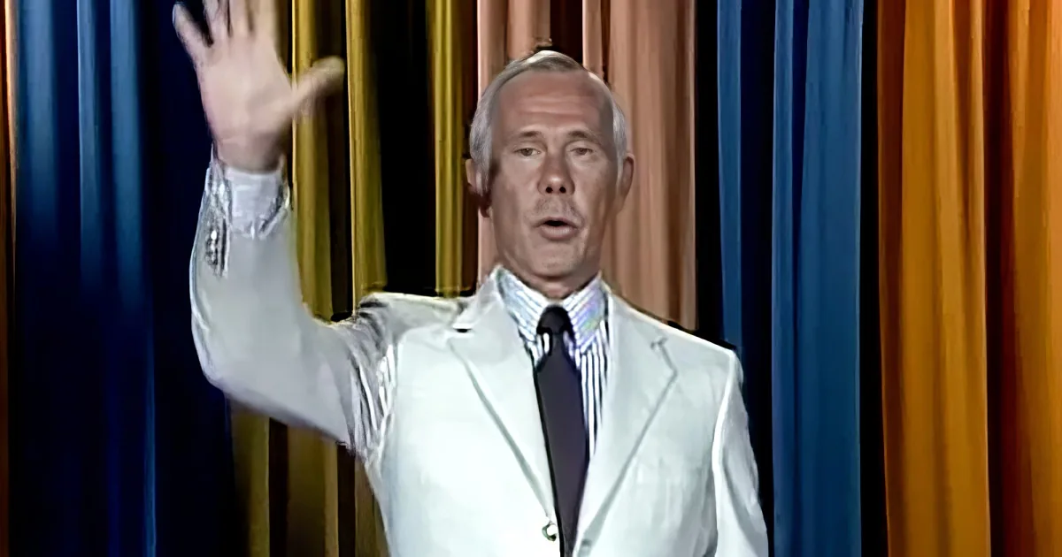 A man in a white suit raises his hand, standing against a colorful curtain backdrop.