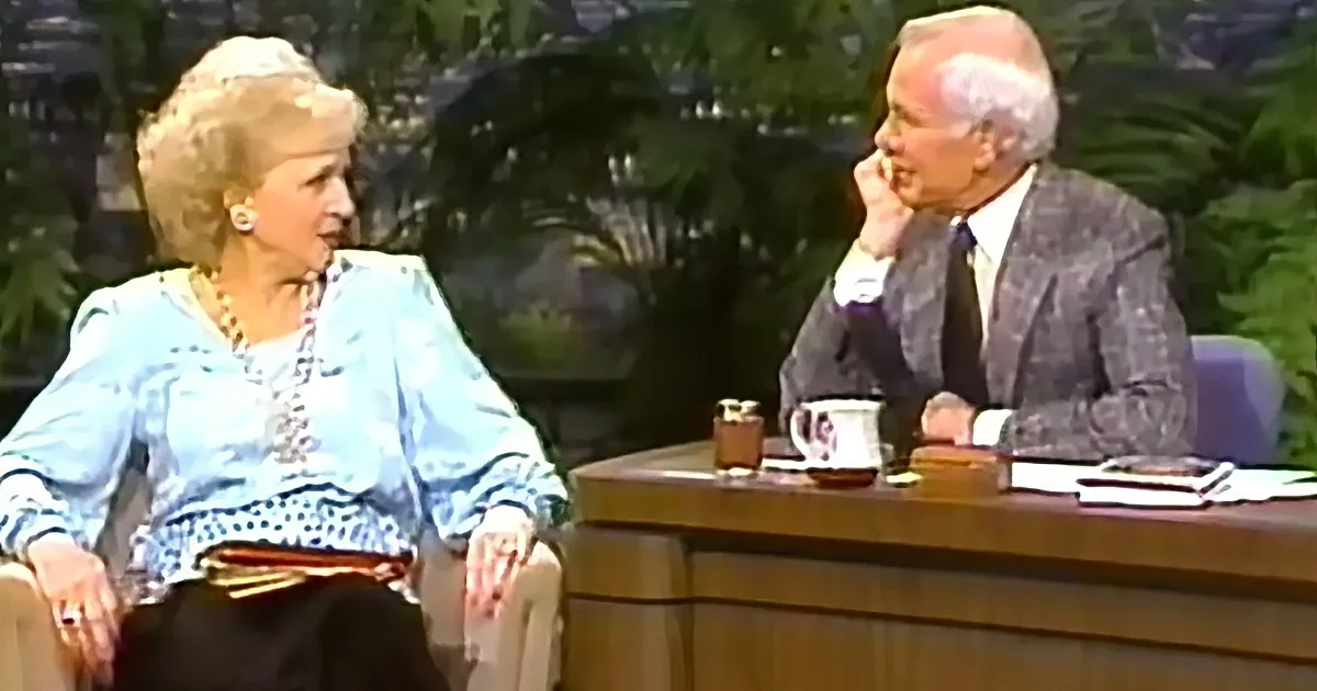 A woman in a blue blouse sits on a chair, engaged in conversation with a man in a suit at a desk.