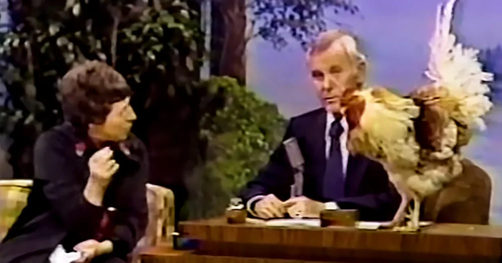 A man in a suit and tie is seated at a desk, holding a microphone, with a person in a costume behind him.