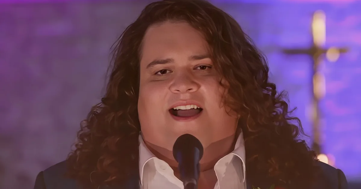 A person with curly hair sings passionately in front of a purple background with a cross.