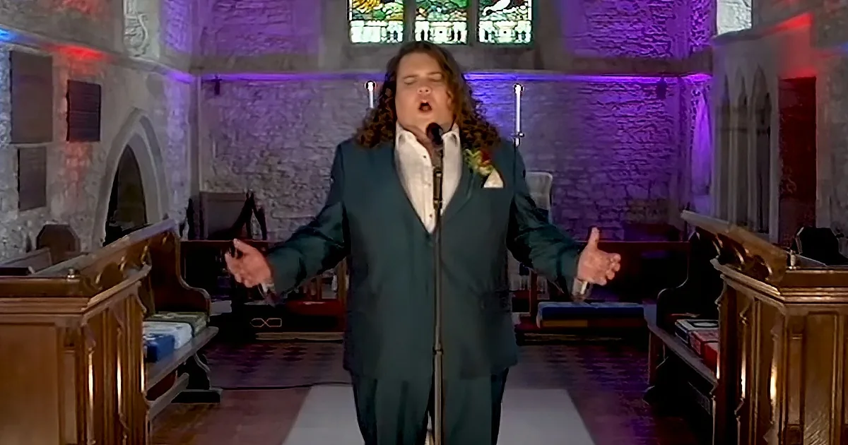 A person in a teal suit sings passionately in a church with stained glass and wooden pews.