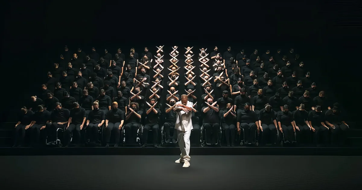 A person in white stands on stage, facing a large group of people in black, who are seated and making X shapes with their...