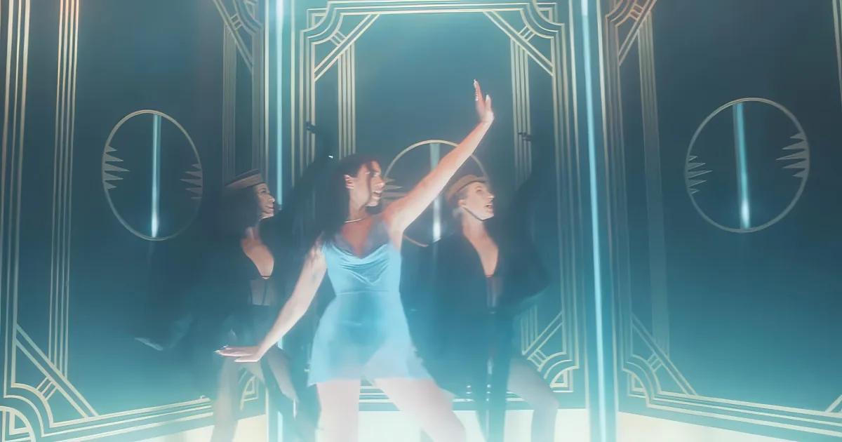 A dancer in a blue dress raises her arm, surrounded by other dancers in a dark, illuminated setting.