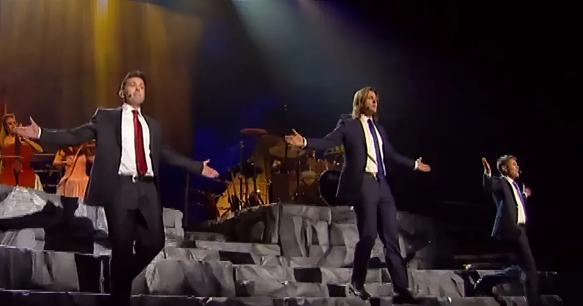 Two men in suits with arms outstretched on stage, performing in front of an orchestra.