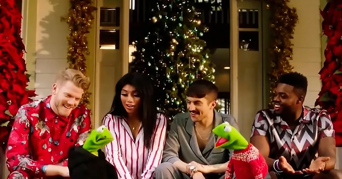 Four people and Kermit the Frog in festive attire, smiling in front of a Christmas tree.