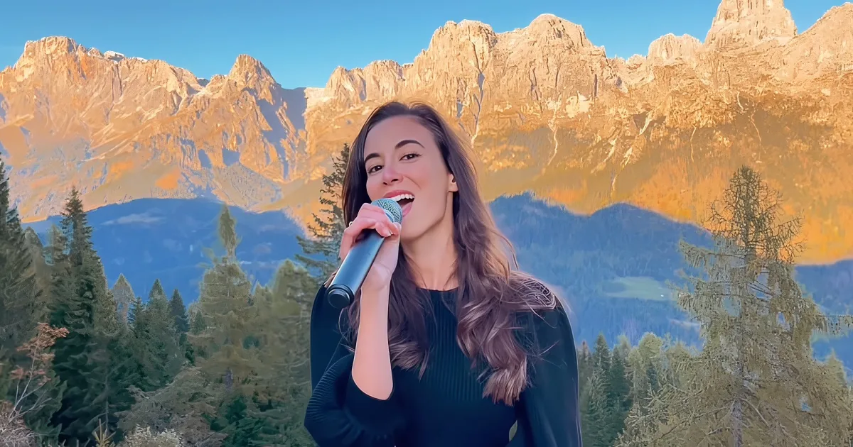 A woman sings into a microphone against a backdrop of majestic, sunlit mountains and evergreen trees.