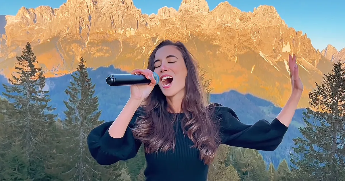 A woman sings passionately into a microphone against a backdrop of majestic mountains and trees.