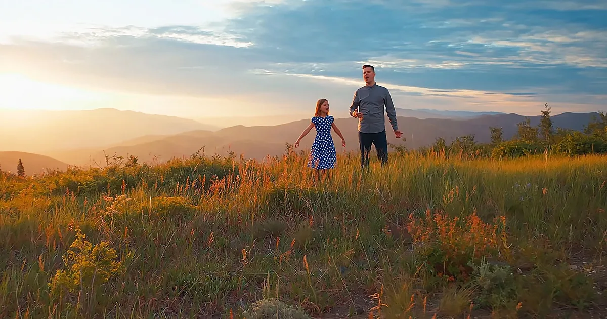 A man and a girl stand in a grassy field, singing under a sunset sky with mountains in the background.