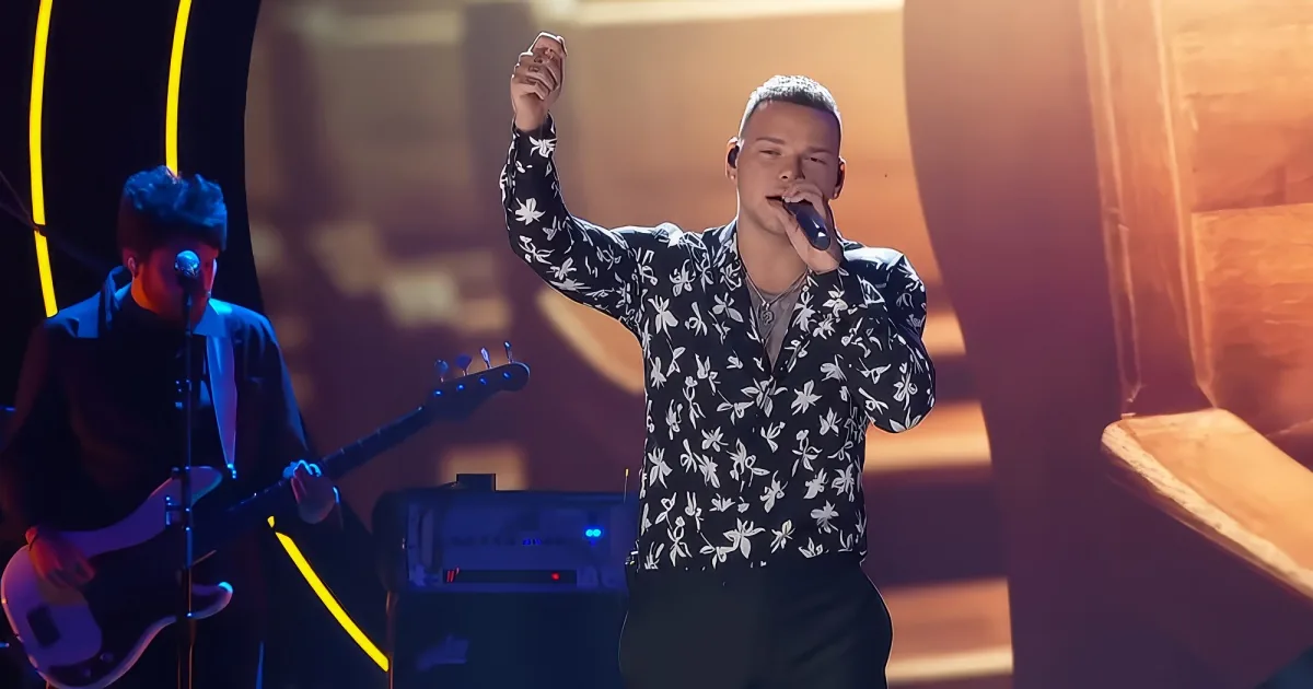 A man in a floral shirt sings into a microphone, raising his other hand, while another musician plays guitar in the...