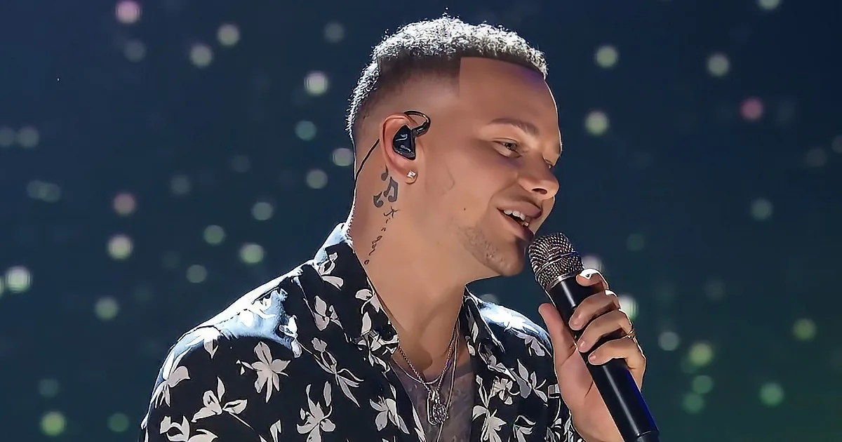 A man with short hair and earphones, wearing a floral shirt, sings into a microphone on stage.