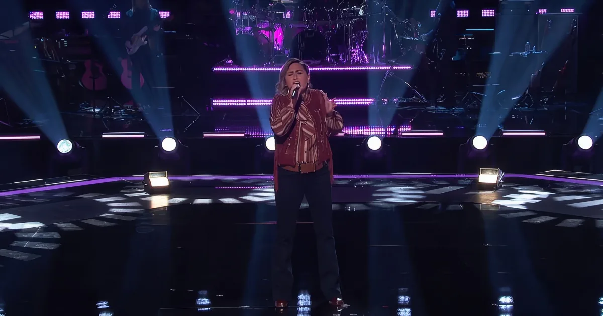 A woman in a red jacket sings on stage with bright spotlights and a band in the background.