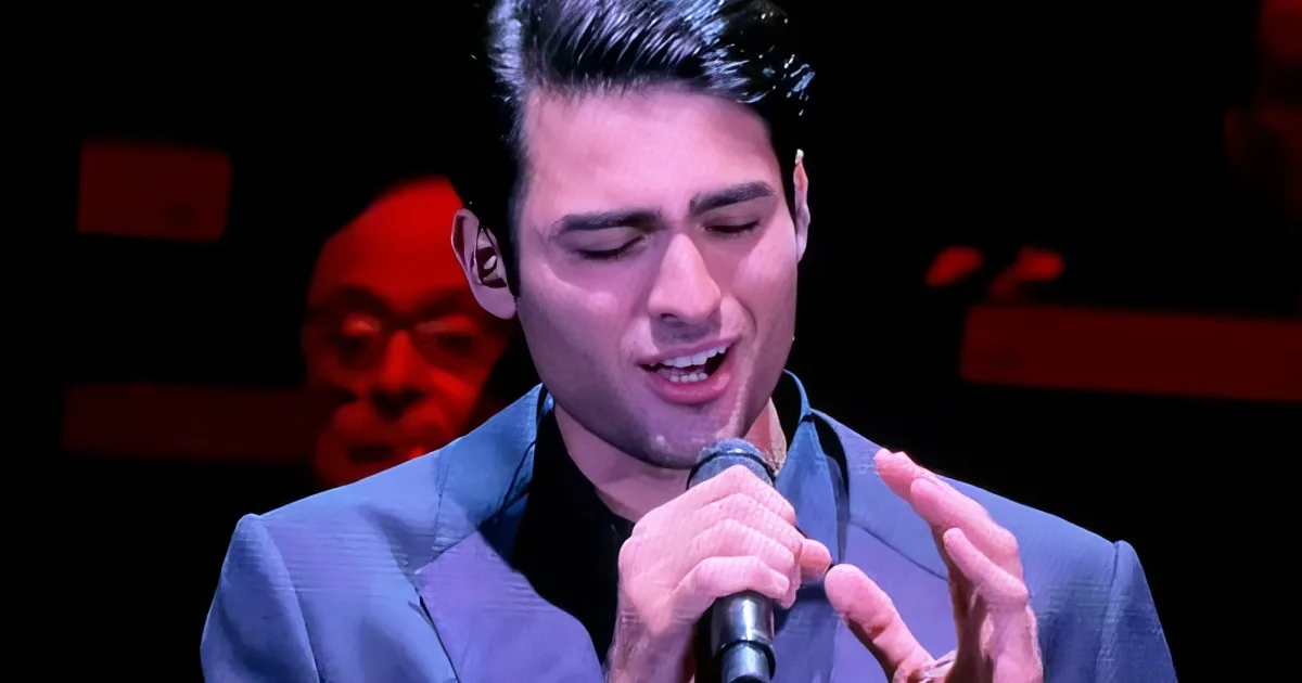 A man in a suit sings passionately into a microphone, his hands clasped together.