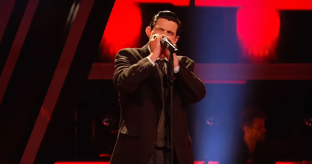 A man in a suit sings passionately into a microphone on a stage with red lighting.