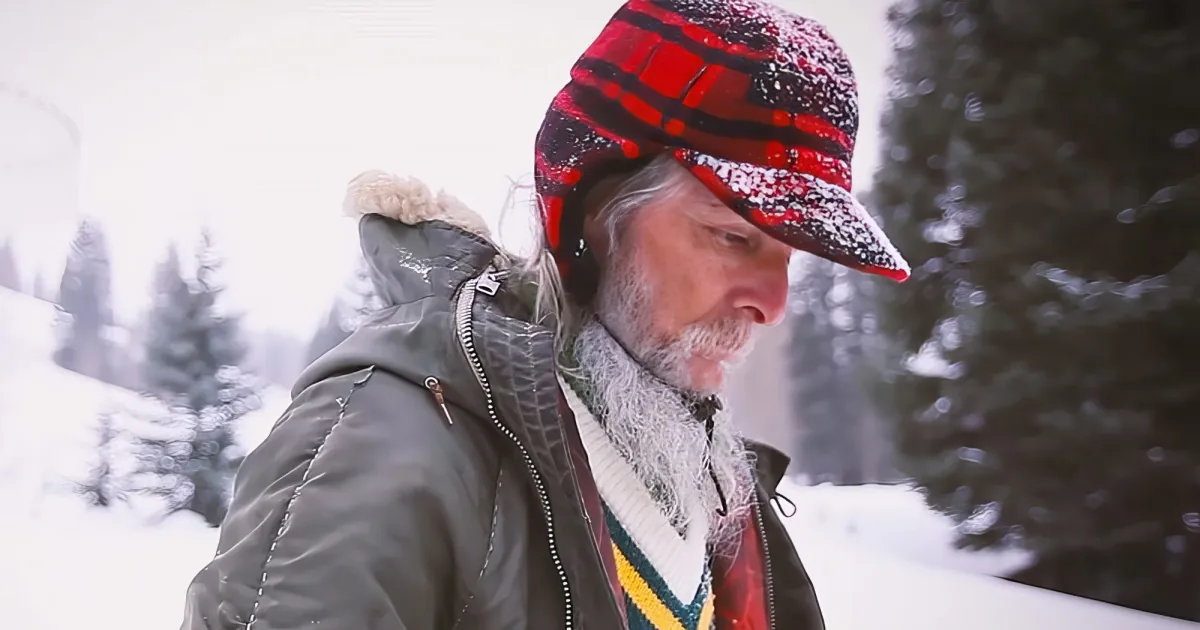 An older man with a red plaid hat and gray beard stands in a snowy forest, looking down.