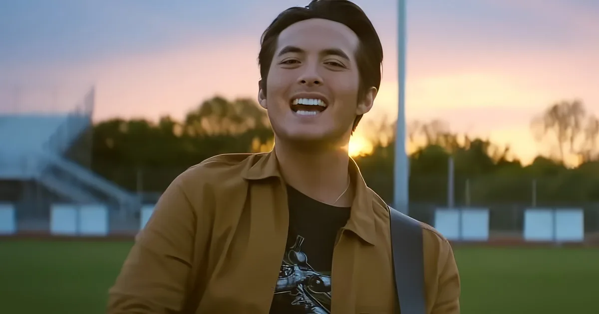 A smiling person in a brown jacket stands outdoors with a sunset and trees in the background.