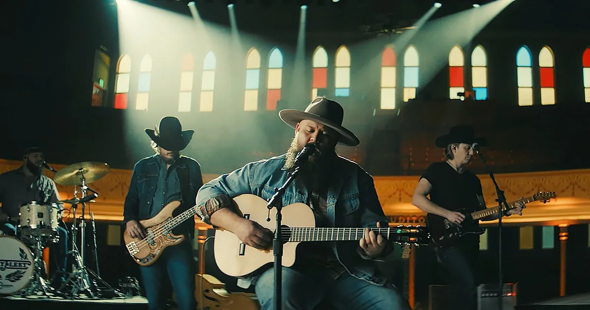 A bearded man in a hat plays guitar on stage with a band in a church setting.