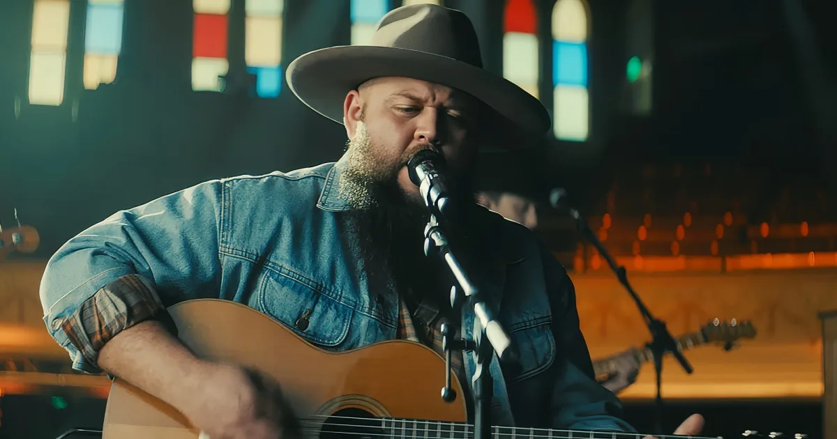 A man with a beard and hat plays guitar and sings into a microphone in a church setting.