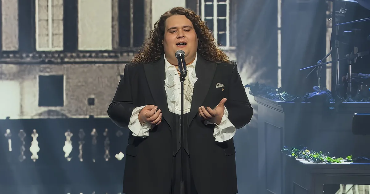 A man in a black suit and white ruffled shirt sings into a microphone on stage.
