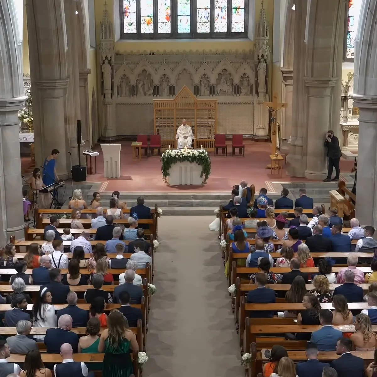 A priest stands at the altar in a church, surrounded by seated guests during a wedding ceremony.