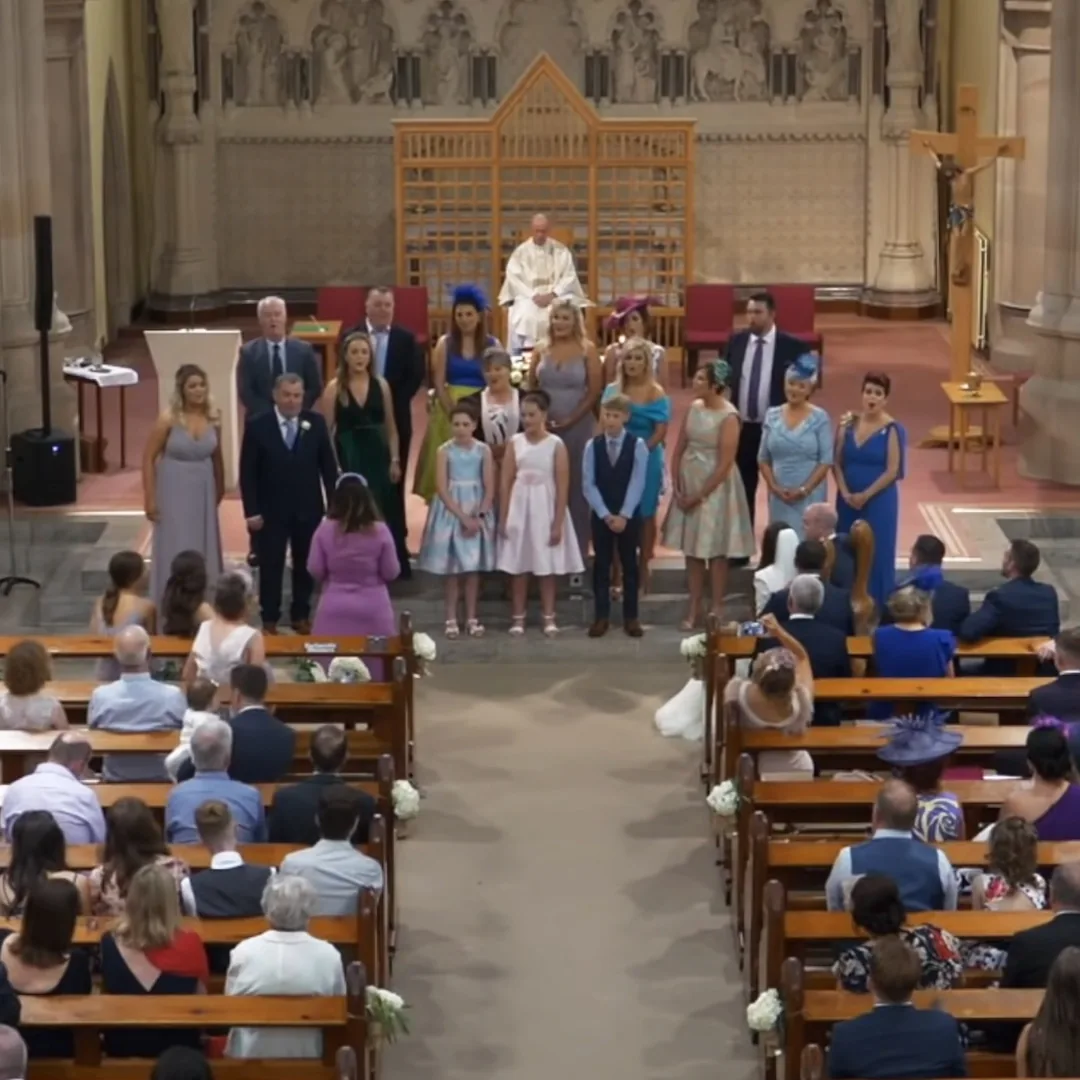 A wedding ceremony in a church with a priest, bridesmaids, and flower girls standing in front of the congregation.