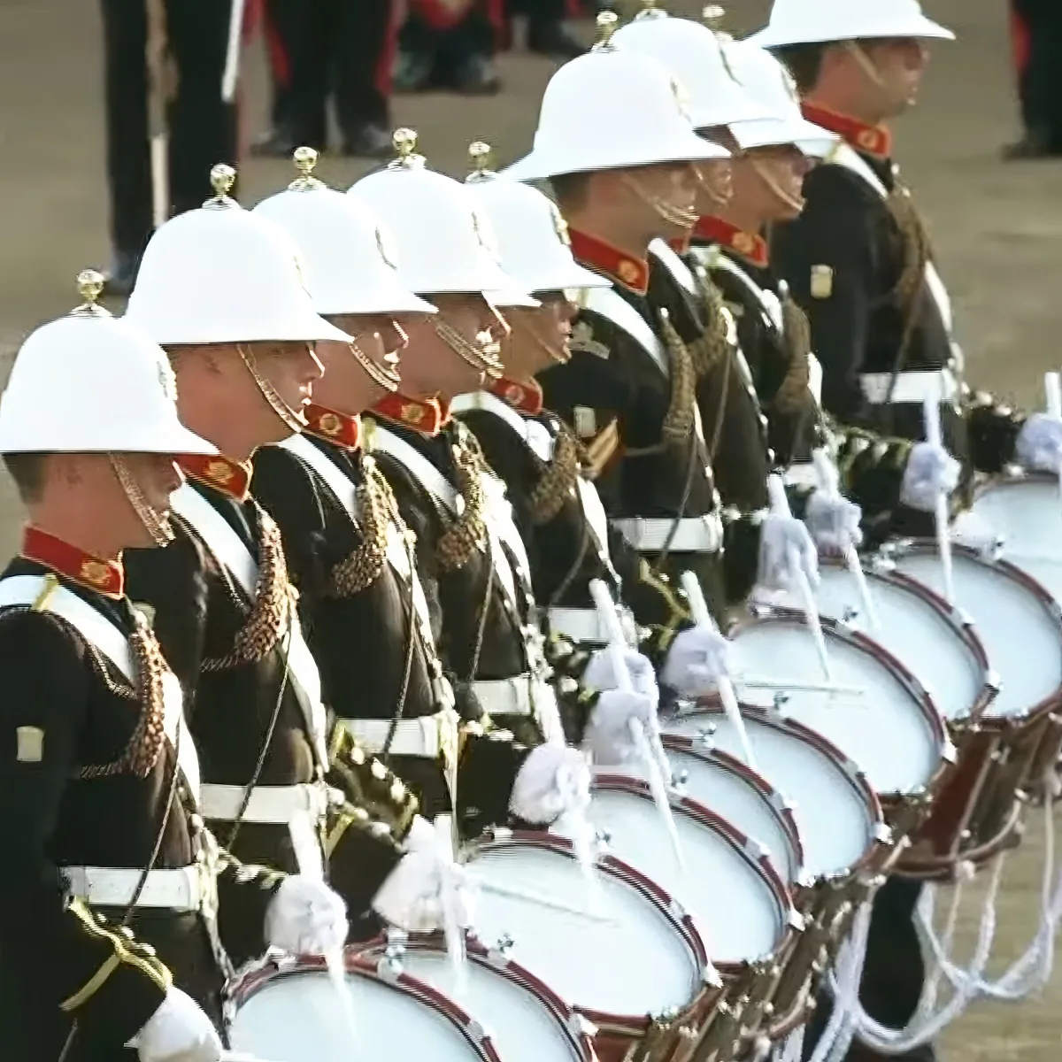 A row of uniformed drummers in white helmets and black uniforms playing drums in a parade.