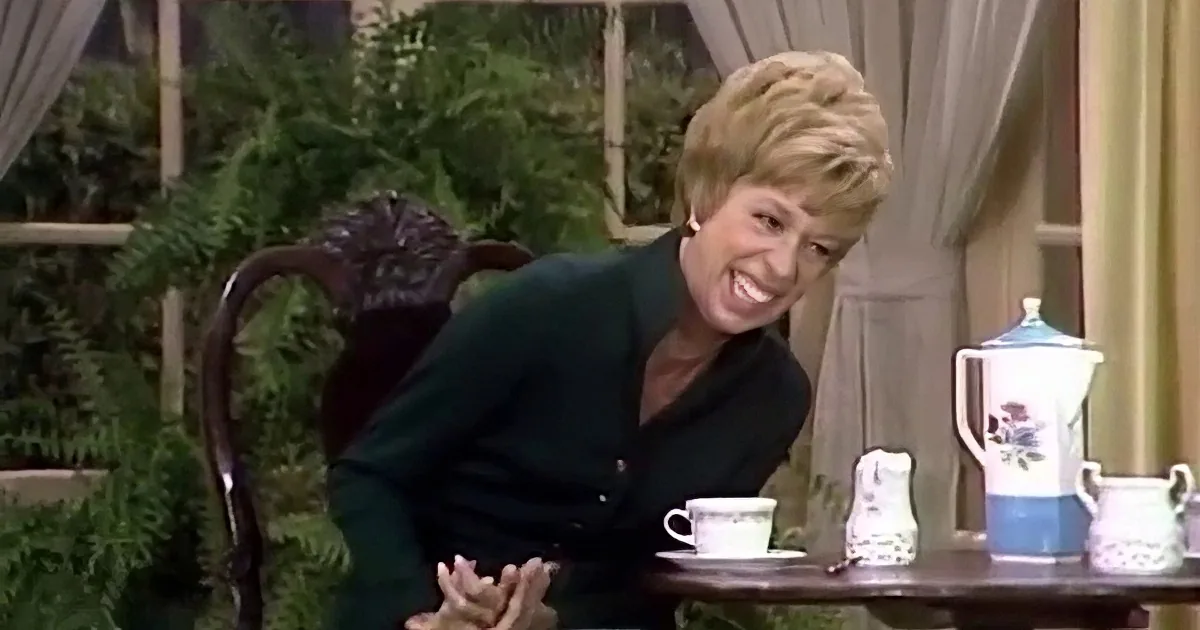 A smiling woman in a dark green cardigan sits at a table with a teapot and cups, surrounded by greenery and curtains.