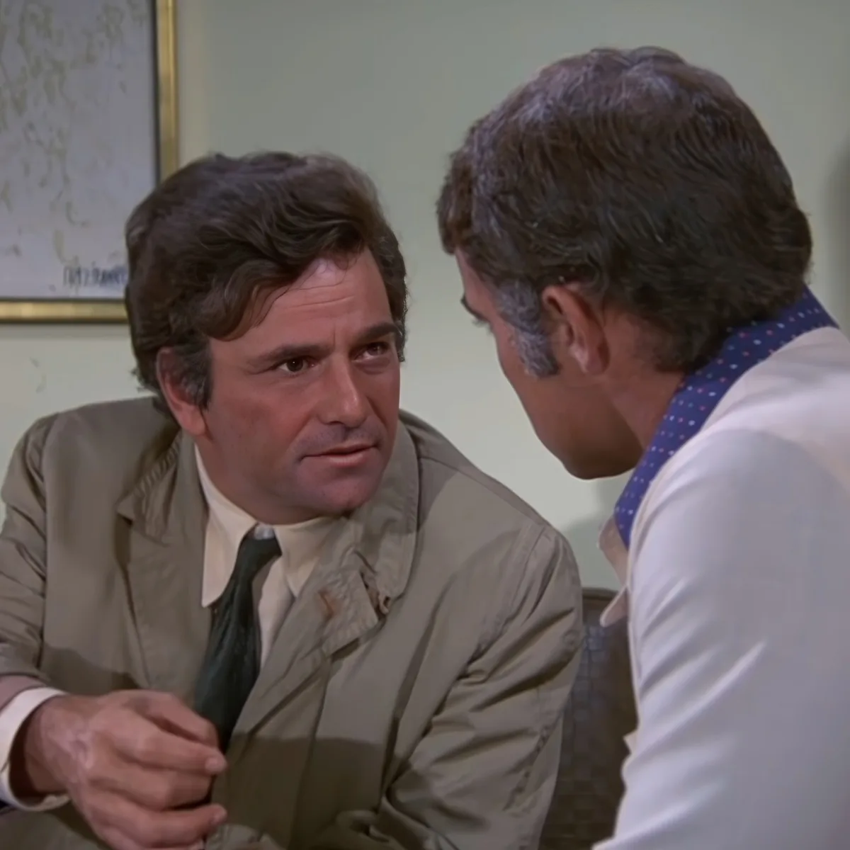 A man in a beige suit adjusts his tie while another man in a white shirt and blue tie looks on.