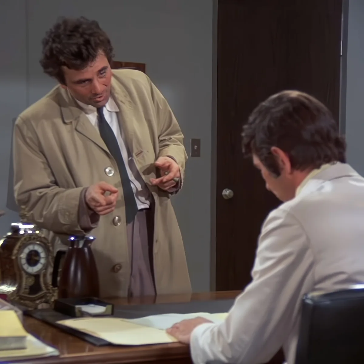 A man in a tan coat gestures while speaking to another man in a white lab coat at a desk.