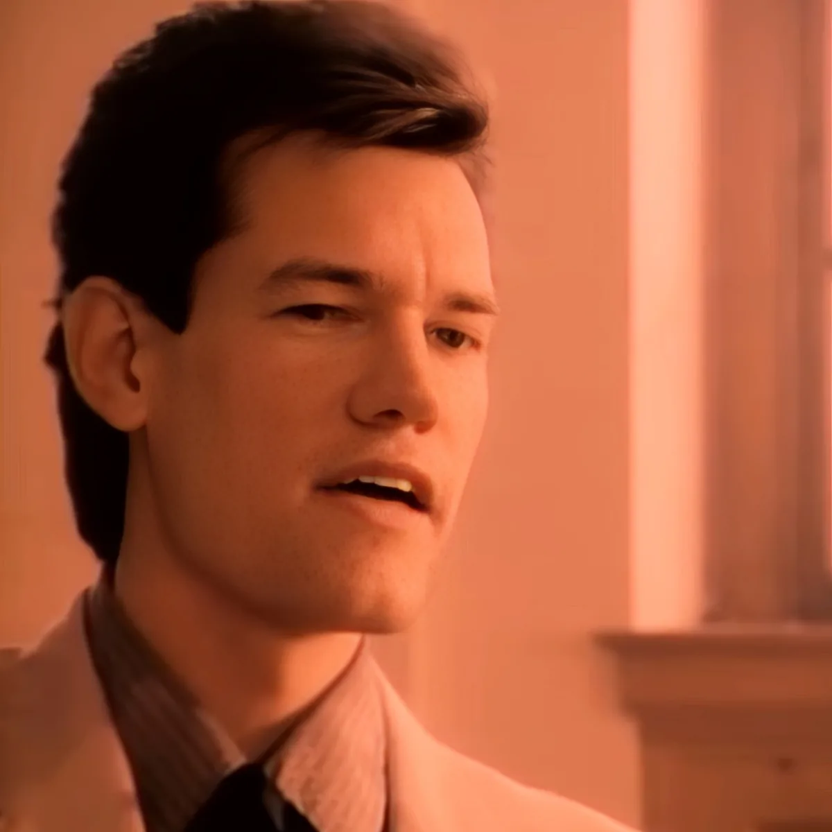 A man with dark hair, wearing a white shirt and tie, appears to be speaking or reacting to something.